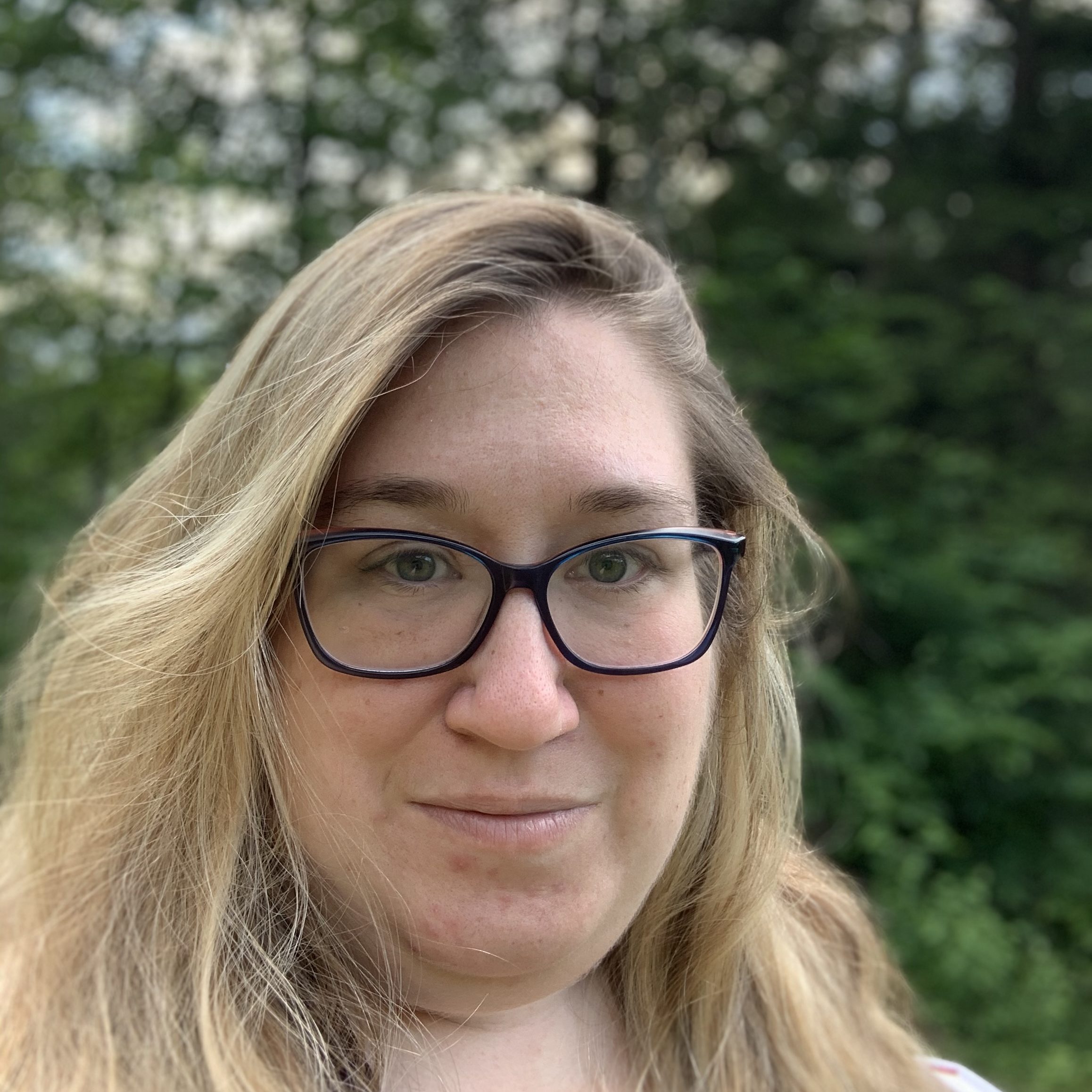 Image of a white woman with long blonde hair, wearing glasses and has a soft smile. There are trees blurred in the background.